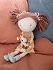 A plush doll sitting on a textured surface, holding a small monkey plush toy. Produktbild-Vorschau
