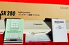 A knitting machine, its instruction manuals, and punch cards are laid out on a red surface in front of its original packaging. Produktbild-Vorschau