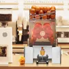 A commercial orange juicer is shown in a cafe or juice bar setting, with a coffee grinder and menu in the background. Anteprima dell’immagine prodotto