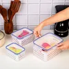 A person's hands are shown placing the lid on a set of three clear plastic food storage containers with white perforated inserts, arranged on a white countertop. Wooden kitchen utensils are visible in a holder in the background, along with a black coffee m Produktbild-Vorschau
