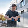 A man is drilling a hole in a piece of wood outdoors, with a modern building in the background. Produktbild-Vorschau