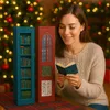 A woman is reading a small book in front of two decorative, vintage-style miniature bookshelves. The background features a decorated Christmas tree with lights. Náhled obrázku produktu