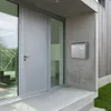 A modern house entrance with a gray door, large glass windows, and a silver mailbox mounted on a concrete wall. Podgląd zdjęcia produktu