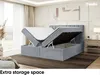 A bedroom scene featuring a gray upholstered bed with its storage compartment open, revealing the mattress and topper. The room has soft lighting and a large window with sheer curtains. Produktbild-Vorschau