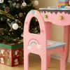 A close-up view of a pink children's vanity table and chair set, decorated with a rainbow and hearts, placed near a Christmas tree and a gift box. Podgląd zdjęcia produktu