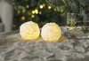 Two snowball-shaped candles are lit and placed on a textured gray tablecloth, with a blurred Christmas tree and lights in the background. Produktbild-Vorschau