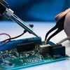 A close-up of a person's hand using tweezers to work on a circuit board, with a soldering iron nearby. Voir les images du produit