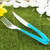 A fork and knife set with blue handles rests on artificial grass, with white stones and a yellow flower in the background. Anteprima dell’immagine prodotto