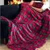 A person is sitting on a couch, covered by a red and green plaid blanket, with a fireplace in the background. Produktbild-Vorschau