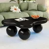 A black, cloud-shaped coffee table with spherical legs sits on a light-colored rug in a living room. A green sofa and a flower-shaped pillow are visible in the background. Voir les images du produit