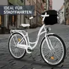 A white city bicycle with a black basket is parked on a cobblestone street in a European town. Produktbild-Vorschau