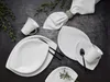 A flat lay of white porcelain dinnerware set on a dark textured surface, with a sprig of greenery. Produktbild-Vorschau