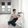 A man is kneeling and using a cordless drill to make a hole in a white wall, with PVC pipes and fittings on the floor. Voir les images du produit