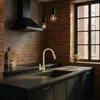 A modern kitchen with brick walls, a black range hood, and black cabinets, featuring a gold faucet over a black sink. Produktbild-Vorschau