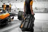 A person in a McLaren racing uniform is pulling a stack of black toolboxes with orange accents next to a Formula 1 car in a garage. Podgląd zdjęcia produktu