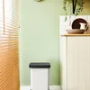 A white trash can with a black lid is placed on the floor next to a light green wall and a cream-colored cabinet. Blinds are visible on the left side of the image. Produktbild-Vorschau