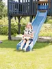 A young girl is sitting on a blue slide, holding a stuffed cow toy. She is in a backyard setting with green grass and trees in the background. Produktbild-Vorschau