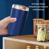Blauer tragbarer Mühle in einer Hand gehalten, neben Glasgläsern mit Gewürzen auf einem Holzregal Náhľad obrázku produktu