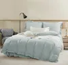 A neatly made bed with light blue bedding, pillows, and a throw blanket, next to a chair and a nightstand with a lamp. Produktbild-Vorschau