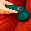 A hand is holding a green fabric shaver over an orange knitted garment. Náhled obrázku produktu