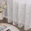A close-up of a white sheer curtain with a gray branch pattern, next to a vase of white flowers and a rug with an open book. Produktbild-Vorschau