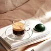 A cup of espresso coffee and a coffee capsule are placed on top of books, with a soft, textured background. Produktbild-Vorschau