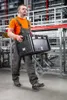 A man in work clothes carries a black plastic crate in a warehouse or factory setting. Produktbild-Vorschau