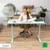 A woman is sitting at a white adjustable desk in a home office setting, writing in a notebook. The desk has dimensions indicated by green lines and text overlays. Produktbild-Vorschau