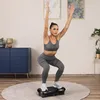 A woman is performing a squat exercise on a vibration plate in a living room. Produktbild-Vorschau