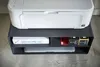 A white printer sits on top of a black multi-tiered shelf unit. The shelf unit contains a stack of paper, three ink cartridges, and a laptop. Produktbild-Vorschau