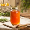 A jar of honey sits on a wooden board, with pears in a basket in the background. Podgląd zdjęcia produktu
