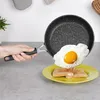 A hand is holding a frying pan with a fried egg being poured onto toast on a plate. Náhled obrázku produktu