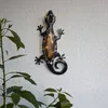 A solar-powered gecko-shaped wall decoration is mounted on a textured white wall, with green plants in the foreground. Produktbild-Vorschau