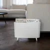 A white magazine rack with magazines inside, placed on a dark wooden floor next to a sofa. Podgląd zdjęcia produktu