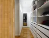 A hallway with a wooden door on the left and a closet with white shelves and drawers on the right. A full-length mirror is visible in the distance. Podgląd zdjęcia produktu