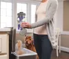 A woman is emptying a vacuum cleaner canister into a trash can, with a dog watching in the background. The scene appears to be in a home kitchen or dining area. Podgląd zdjęcia produktu