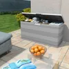 A grey storage box is open and filled with towels and other items, placed on a patio next to a lounge chair. A glass coffee table with a bowl of oranges and an open book is in the foreground. Produktbild-Vorschau
