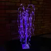 A decorative illuminated willow tree with purple lights stands on a dark surface in front of a brick wall. Podgląd zdjęcia produktu
