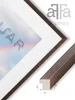 A close-up view of a framed picture with a wooden frame and a white mat. A sample of the frame material is shown in the foreground with dimensions. Produktbild-Vorschau