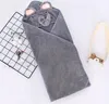 A grey hooded towel with an elephant face design is laid out on a white surface, with a black wire grid and some decorative items to the left, and a small wooden camera to the right. Produktbild-Vorschau