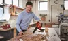 A man is using a red and black leaf blower in a workshop to clear wood shavings from a large wooden table. Produktbild-Vorschau
