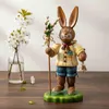 Ein fröhlicher Hase in traditioneller Kleidung hält einen Stab mit Blumen in der einen Hand und eine Glocke in der anderen während er auf einem grünen Sockel steht und freundlich lächelt Produktbild-Vorschau