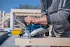 A person is using a cordless circular saw to cut a piece of wood at a construction site. Podgląd zdjęcia produktu