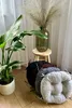 A collection of round seat cushions in various colors are arranged on a wooden floor next to a potted plant and a stool with a vase of dried flowers. Produktbild-Vorschau