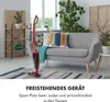 A red cordless vacuum cleaner is displayed upright in a living room setting, standing on a rug in front of a grey sofa. The sofa has several decorative pillows. A wooden shelf unit with plants and decorative items is visible in the background. Produktbild-Vorschau