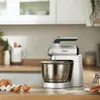 A silver Kenwood hand mixer is shown in a kitchen setting, with eggs in a carton and cracked eggs on the countertop. Produktbild-Vorschau