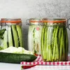 Three glass jars filled with pickled vegetables, including cucumbers, cabbage, and asparagus, are displayed on a red and white checkered cloth. A whole cucumber and a red chili pepper are in the foreground. Anteprima dell’immagine prodotto