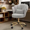 A light gray velvet office chair with a scalloped back and a small lumbar pillow is shown in a home office setting. The chair has a gold base with wheels and is placed on a striped carpet. Behind the chair, a wooden shelf unit is visible with various items Produktbild-Vorschau