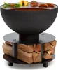 A black metal fire pit with a grill grate on top, holding food items, and a wood storage base. Produktbild-Vorschau