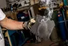A person's arm and hand wearing a glove are holding a black foam pad that is emitting smoke. The background appears to be a workshop or garage with tools and equipment visible. Produktbild-Vorschau
